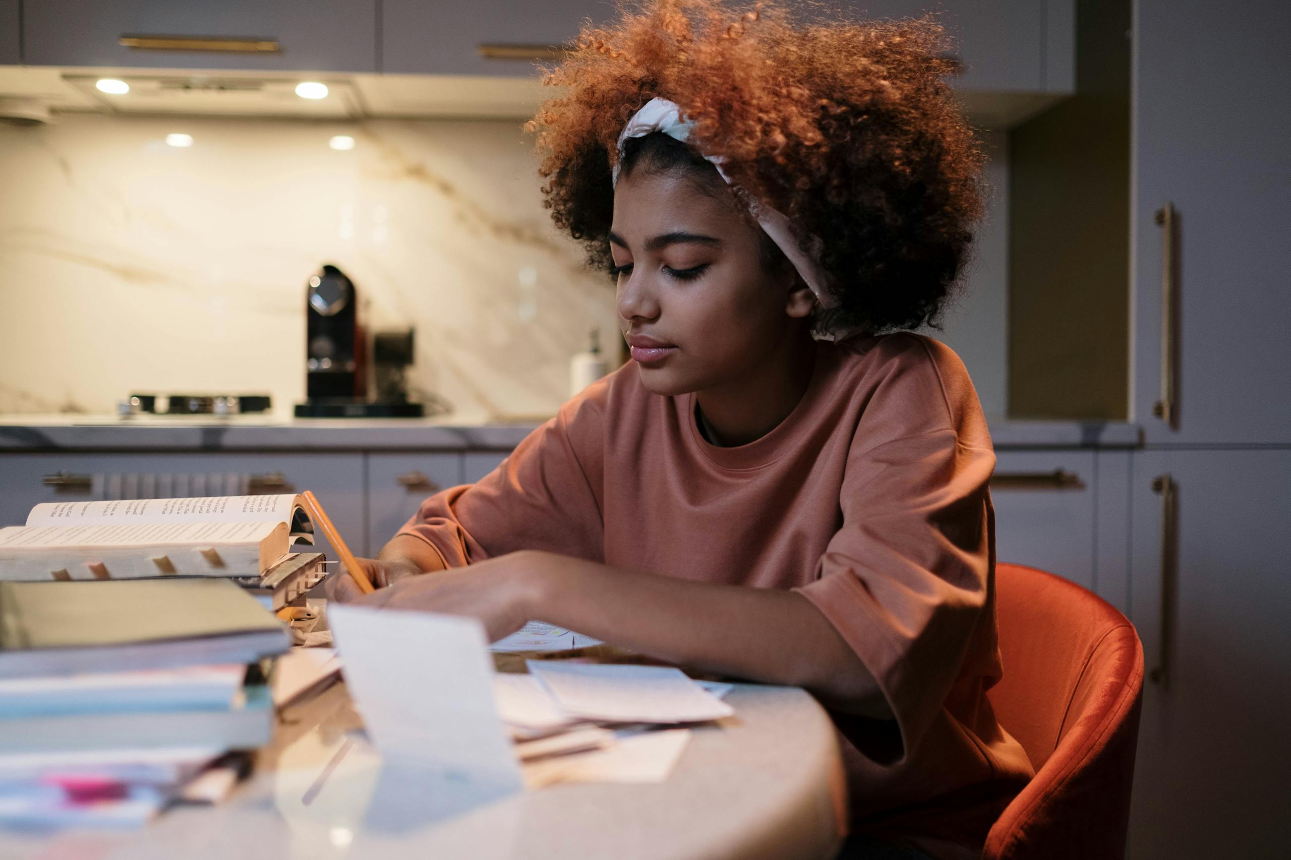 Young woman with curly hair studying at a kitchen table, surrounded by books and notes.